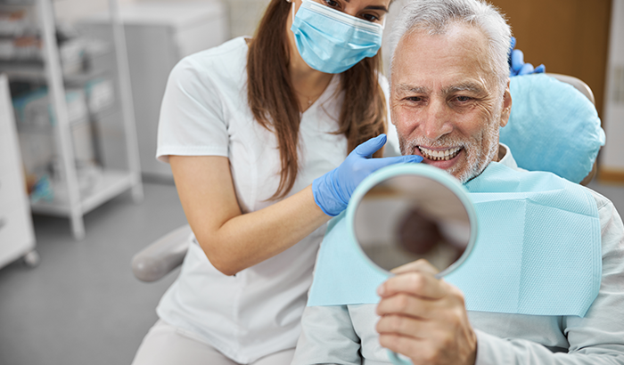 Older man sitting in a dental chair looking in a handheld mirror at his new dental crowns from Dental Care of Burlington in Burlington, MA