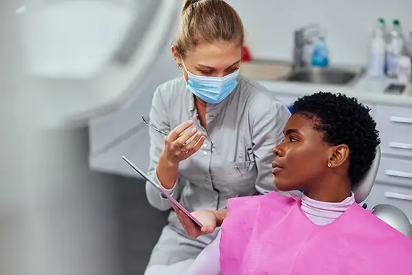 A dentist wearing a mask consulting a female patient, using a tablet to discuss her thyroid disease during an examination.