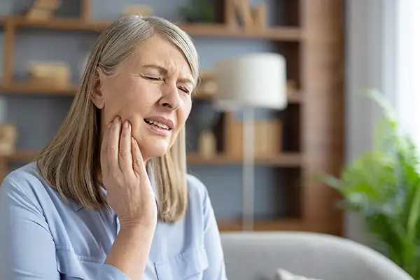 Elderly woman sitting at home, holding her jaw and grimacing in pain, indicating a tooth sensitivity.