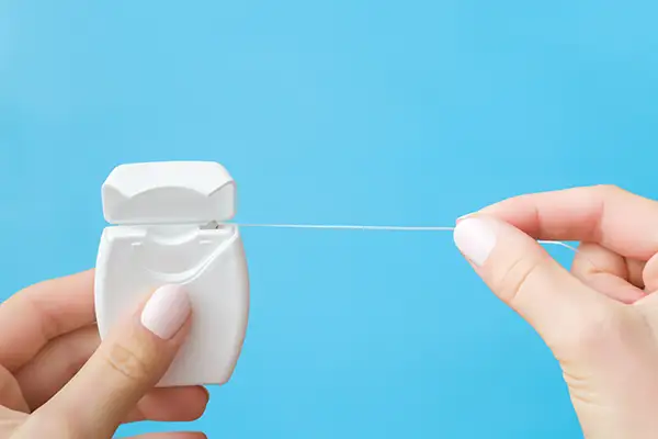 Close-up of hands pulling dental floss from a dispenser against a bright blue background, emphasizing the importance of flossing.