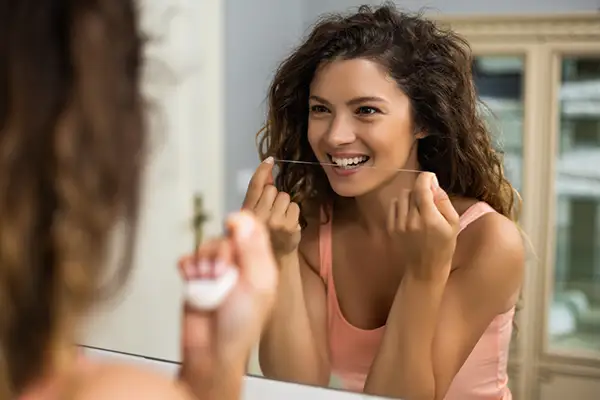 Woman smiling while flossing her teeth in front of a mirror, showcasing an effective daily oral care routine.