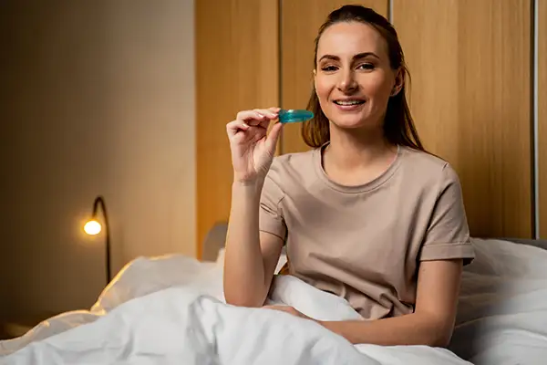 Woman sitting in bed holding a blue mouth guard, preparing for a restful sleep with teeth grinding protection.